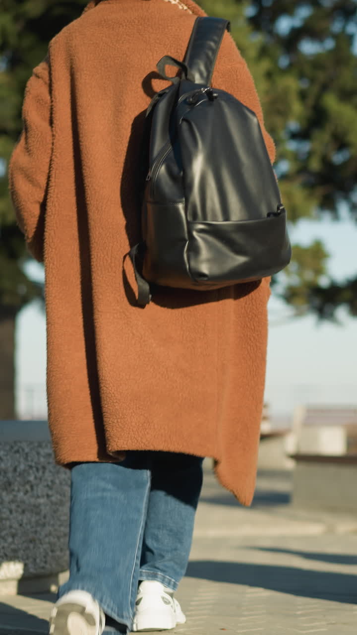 женщина в коричневом пальто, синих джинсах и белых ботинках идет одна по городу с черным рюкзаком. ее поза и медленный ход предполагают печаль или размышление