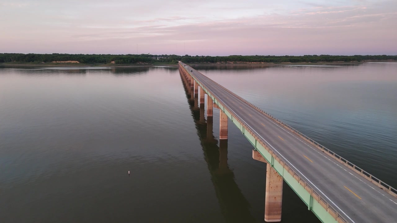 Riprese aeree con drone del Mile Long Bridge al lago Saylorville, Iowa, che mostrano la passerella pedonale in legno sopraelevata che si estende su acque calme e riflettenti con un cielo crepuscolare rosa e nuvoloso.