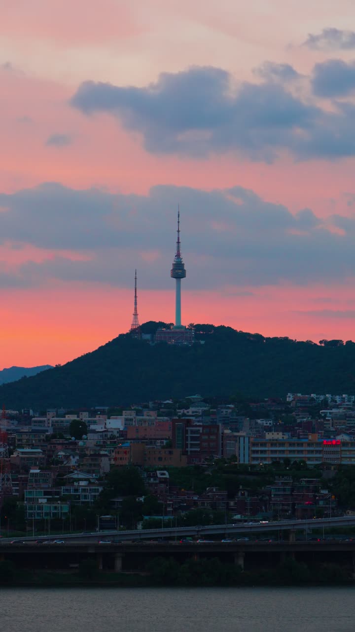 Telephoto vertical close-up shows N Seoul Tower rising over vibrant city buildings, dramatic pink and orange sunset clouds filling the sky above Han River