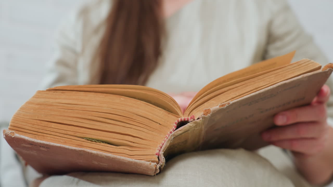 close view of old book resting on fabric surface, partial view of woman flipping thick yellowed pages, visible textured spine with aged details, soft indoor setting, nostalgic and vintage atmosphere