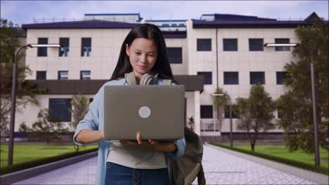 una estudiante adolescente asiática con una mochila usa una computadora portátil y celebra mientras está de pie frente a un edificio escolar