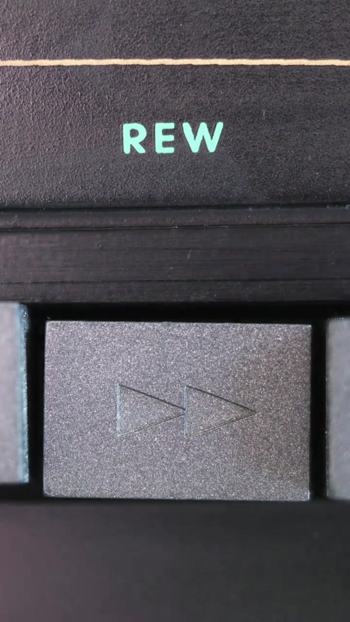 Vertical close up of gray rewind button being pressed below turquoise REW text on black panel