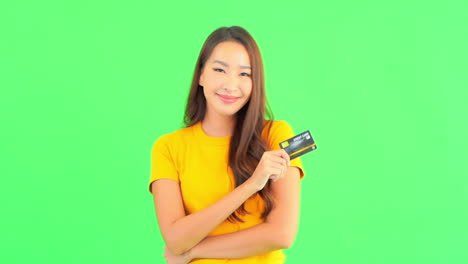 Excited-young-Thai-woman-in-casual-outfit-holding-a-credit-card-in-one-hand-and-smiling-on-green-studio-background