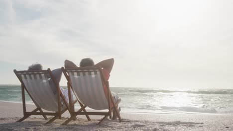 Rückansicht-Eines-älteren-Paares,-Das-Sich-Bei-Sonnenuntergang-Auf-Liegestühlen-Am-Strand-Entspannt