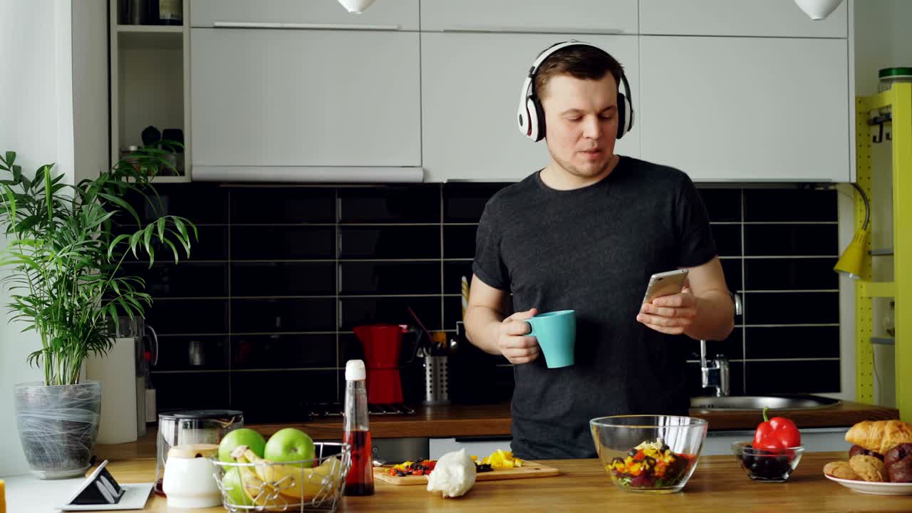un hombre alegre y feliz bailando y cantando en la cocina mientras navega por las redes sociales en su teléfono inteligente en casa por la mañana
