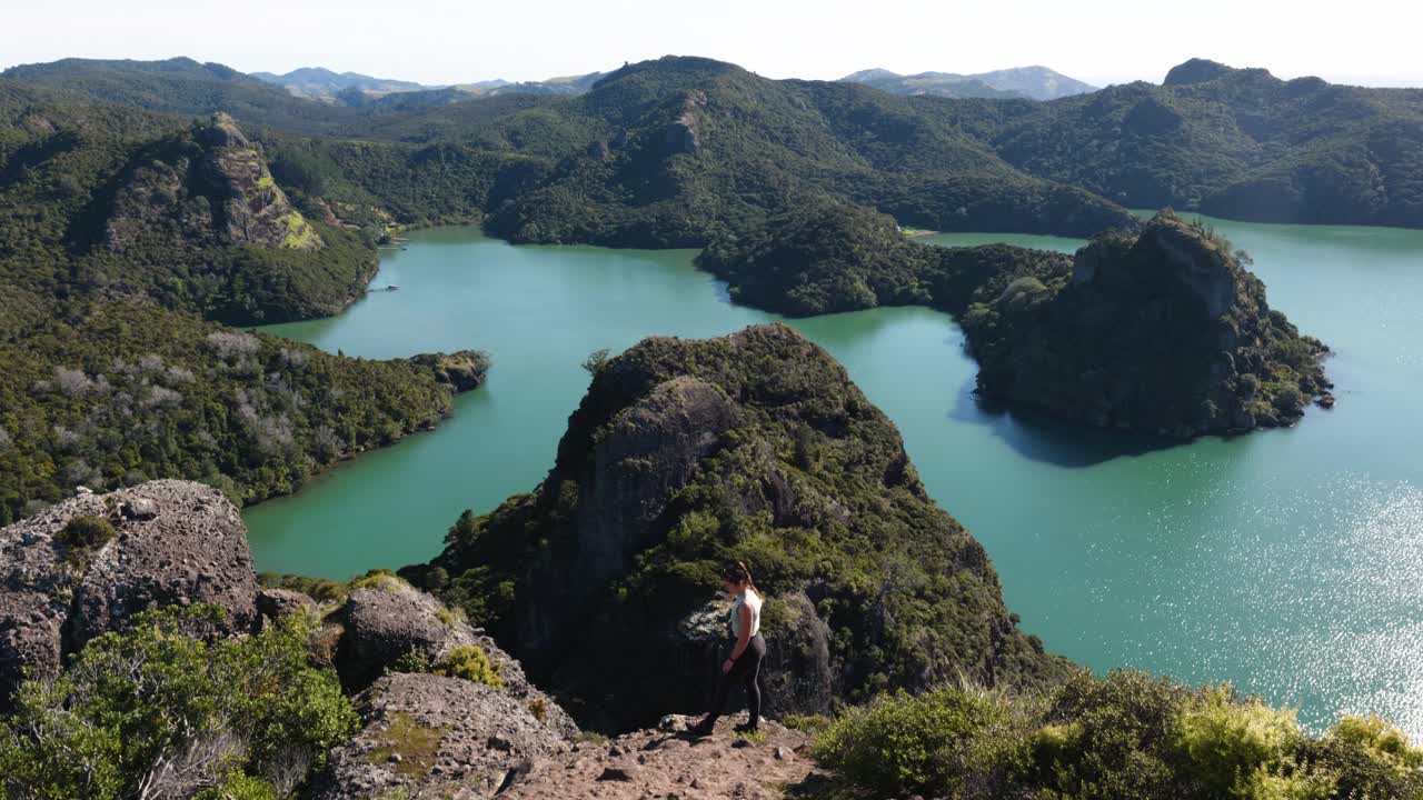 Young caucasian woman standing on a rock looking at a bay with mountains, greenery and turquoise water on a sunny day in Duke's Nose, Northland, New Zealand.