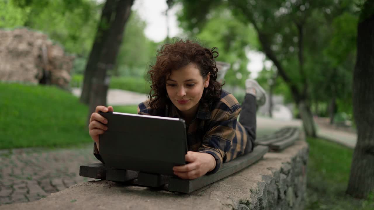 una estudiante feliz con el cabello rizado se acuesta en un banco y mira una tableta durante un descanso entre clases en el parque