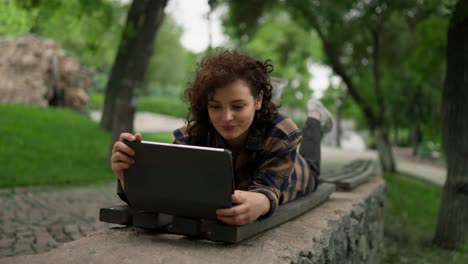 Eine-Glückliche-Studentin-Mit-Lockigem-Haar-Liegt-Auf-Einer-Bank-Und-Schaut-Während-Einer-Unterrichtspause-Im-Park-Auf-Ein-Tablet