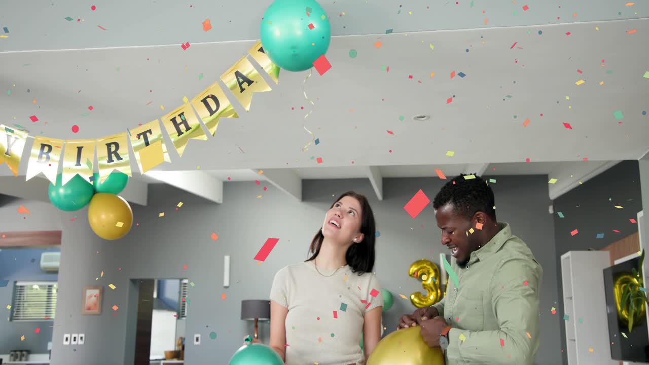 Couple preparing 30th birthday party in living room, confetti bursting, man tying gold balloon