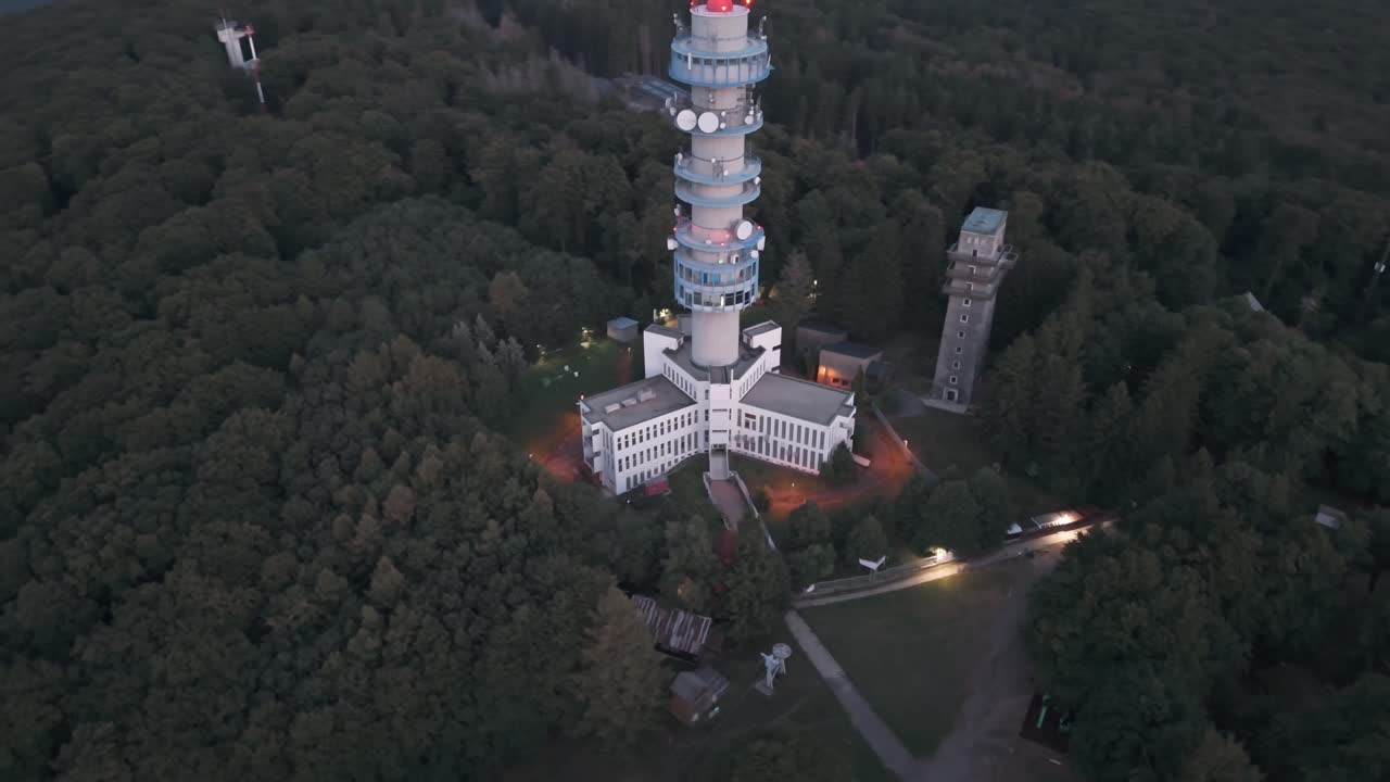 Illuminated base of the Kékestető TV Tower from an orbital drone view at twilight, surrounded by the forests of the Mátra in Hungary