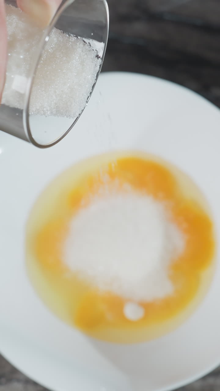 Aerial view of woman preparing to bake, pouring sugar into a bowl of cracked eggs on a dark countertop, whisk placed nearby, indicating upcoming mixing