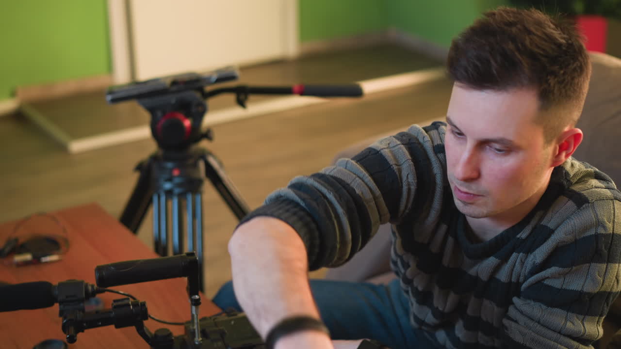 Person adjusting LED video light mounted on tripod. Focus on hands and light setup. Background features soft indoor lighting and couch, with a relaxed home studio atmosphere