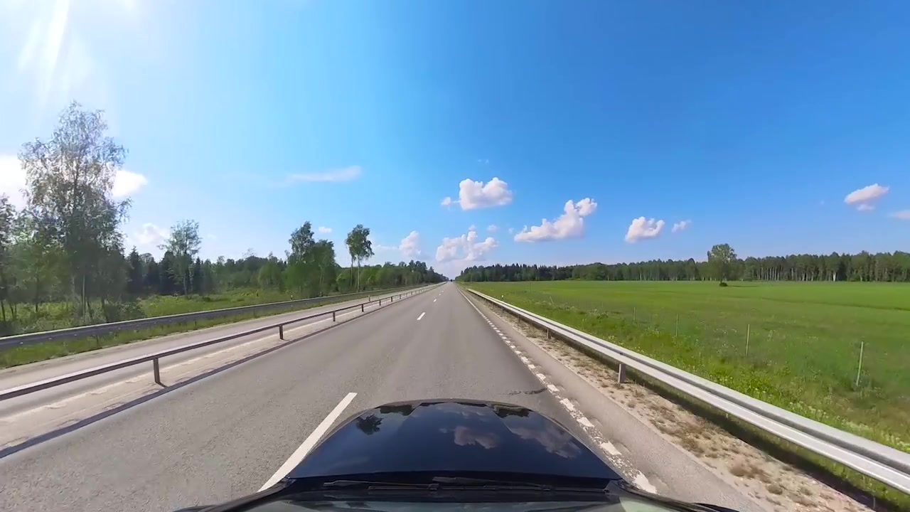 Auto nera che guida in soggettiva su un'autostrada estone durante l'estate con alcune nuvole nel cielo blu. Erba alta verde e alberi visibili ai lati della strada mentre il sole splende. Soggettiva