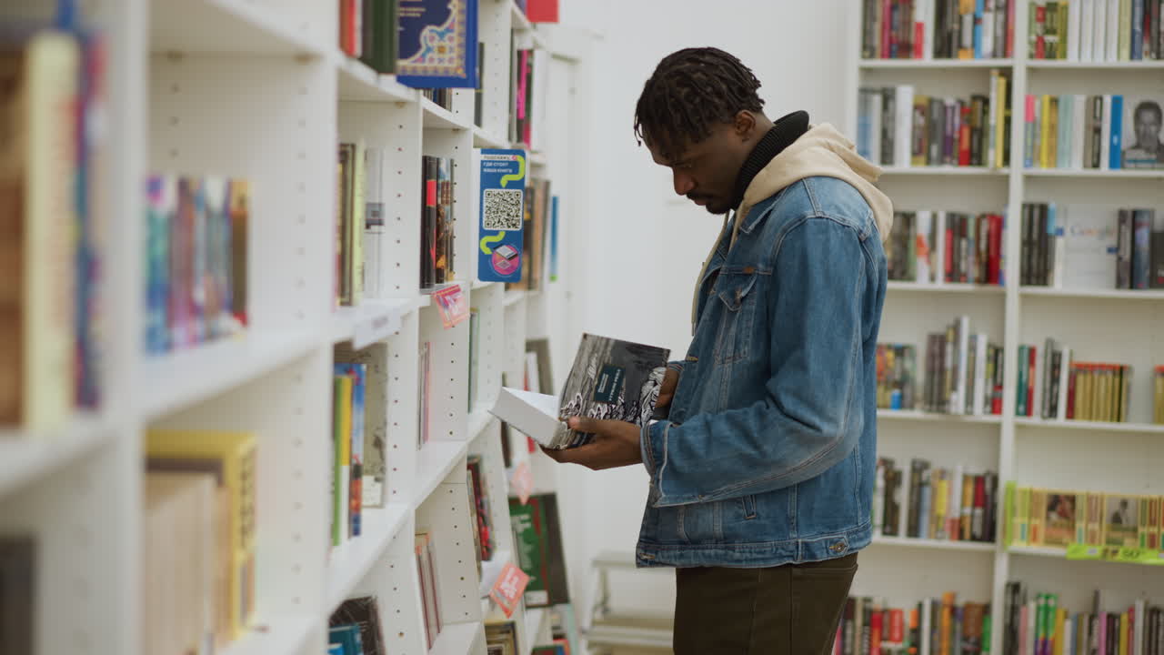 Man in denim jacket browsing bookshelves in bookstore, reaching for book while focused on selection, capturing the joy and concentration of choosing reading material in vibrant environment