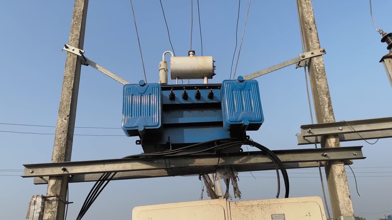 Ripresa orbitale dal basso verso l'alto attorno a un trasformatore di potenza elettrica montato su un palo dell'elettricità, cavi e isolanti visibili mentre la camera ruota su uno sfondo di cielo azzurro limpido