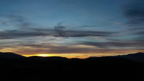 Vuelo-De-Drones-En-Un-Valle-Visualizando-La-Puesta-De-Sol-Con-Todas-Las-Montañas-Oscuras-Un-Cielo-Azul-Amarillo-Y-Naranja-Con-Nubes-Con-Un-Giro-Inclinado-De-La-Cámara-Hacia-Arriba-En-ávila-España