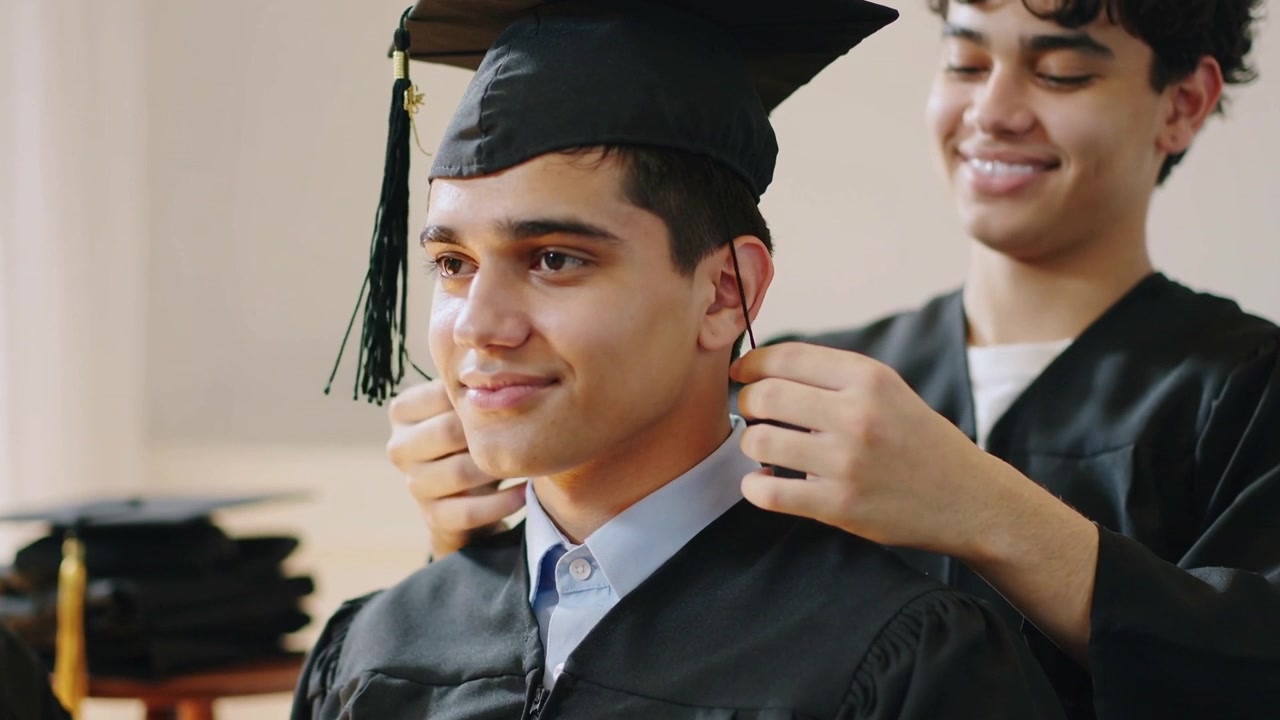 Amico in piedi che si allunga per sistemare il berretto e posizionare il cordino su uno studente in toga nella sala di preparazione per la laurea.
