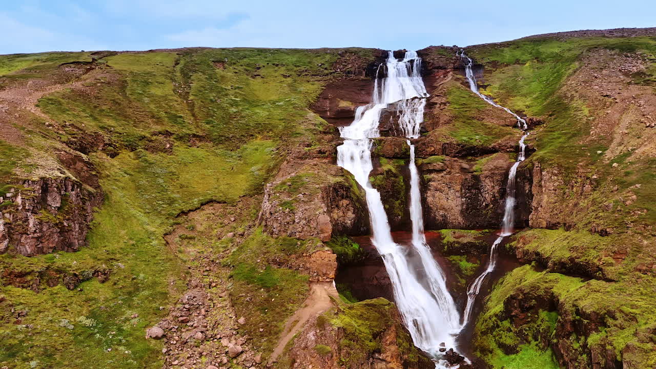 Waterfall splitting into three streams flowing by the bare rock. Going up along the rock covered with mosses with a waterfall on. Iceland nature.