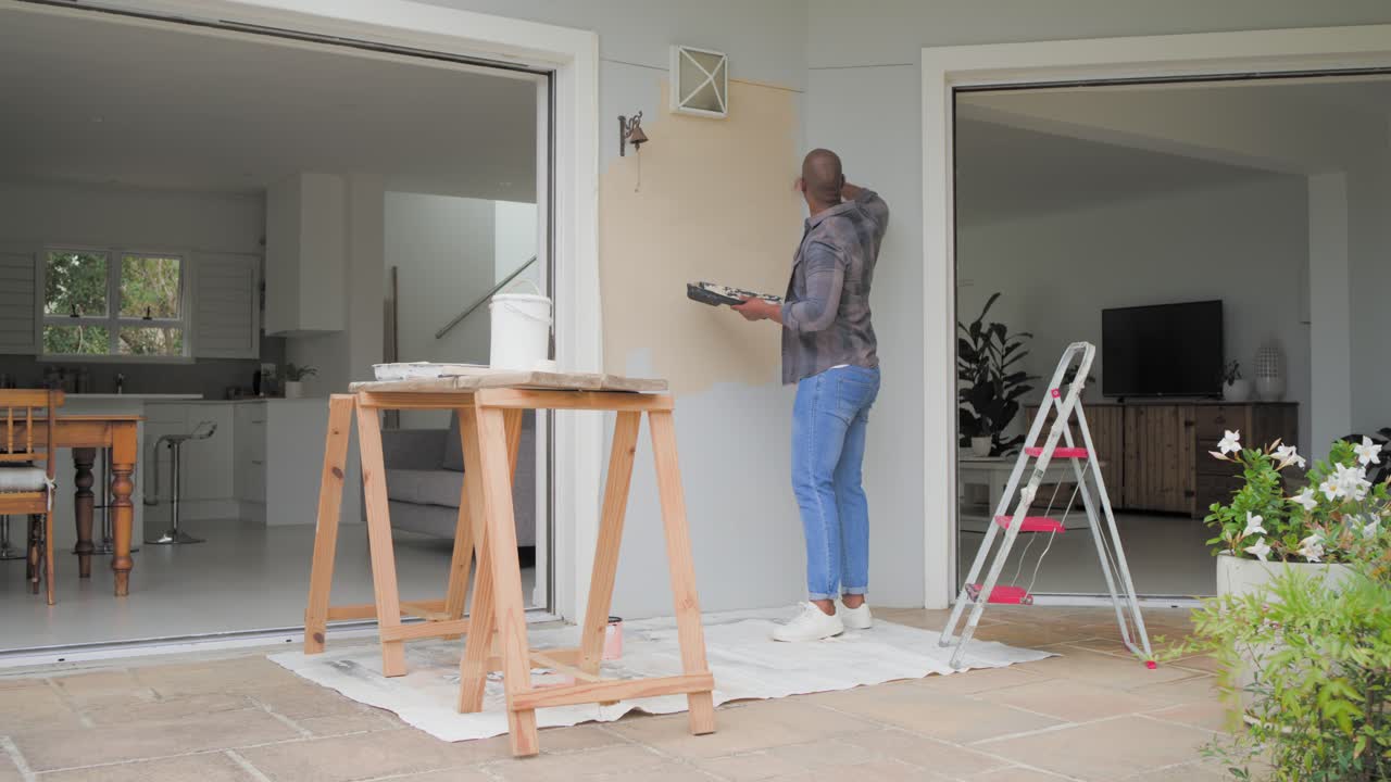Uomo che cammina verso il muro con un vassoio di vernice in mano, mentre dipinge un pannello su un patio coperto per ripararlo, con spazio per il testo.