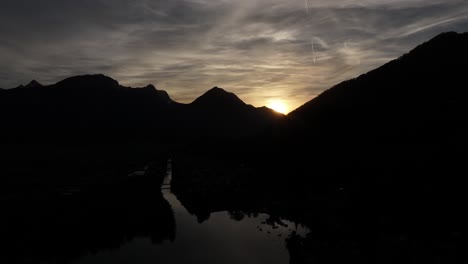 Evening-drone-shot-of-the-Walensee-Lake-and-mountain-ranges-surrounding-Weesen-and-Amden,-Switzerland,-sun-setting-with-dark-mountains-silhouette-and-grey-skies-with-dim-reflection-on-the-lake