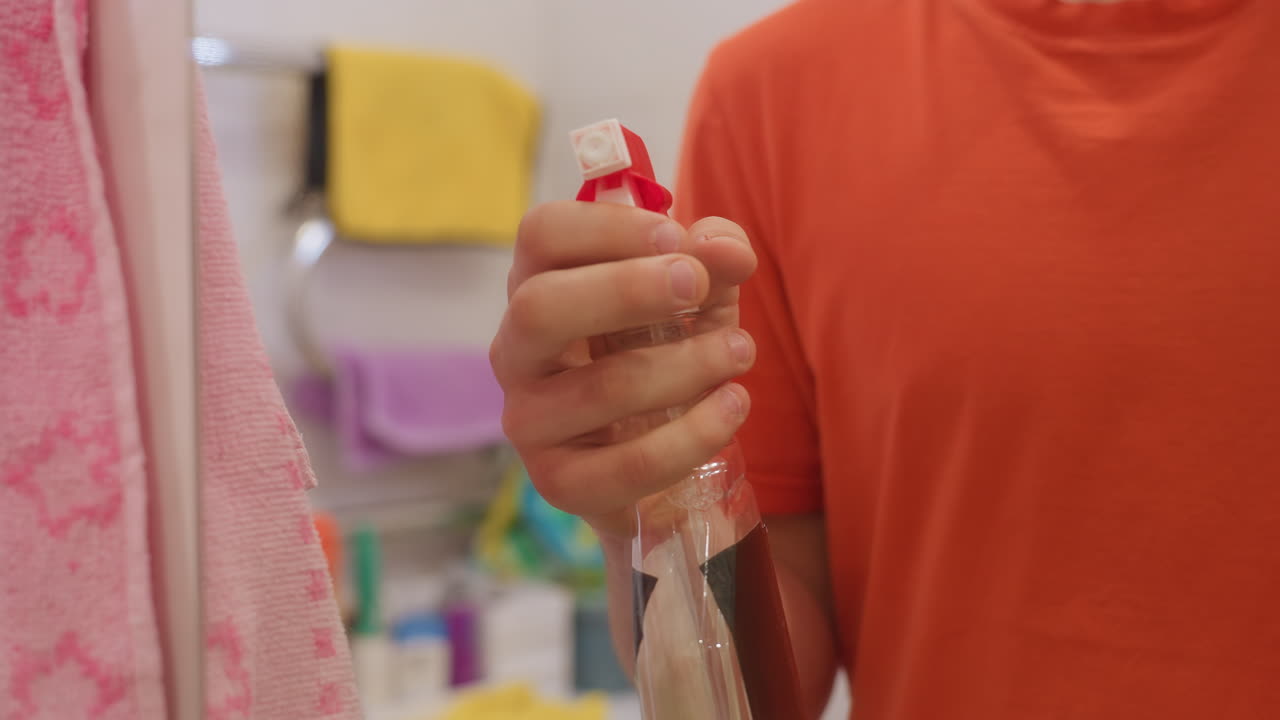 Close up mirror reflection showing man in orange shirt spraying cleaning solution on bathroom surface, holding bottle firmly, focusing on cleanliness, hygiene, sanitation and household routine care