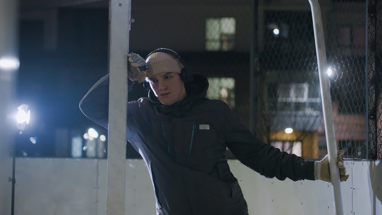 Young man leaning on goal post, wearing gloves, beanie, and headphones under bright night lighting, focusing intently with reflective background of chain-link fence and urban lights