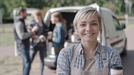 Coffee-Truck-Female-Worker-Looking-At-Camera-Smiling-And-Holding-A-Coffee,-Group-Of-People-Talk-And-Drink-Coffee-In-The-Background