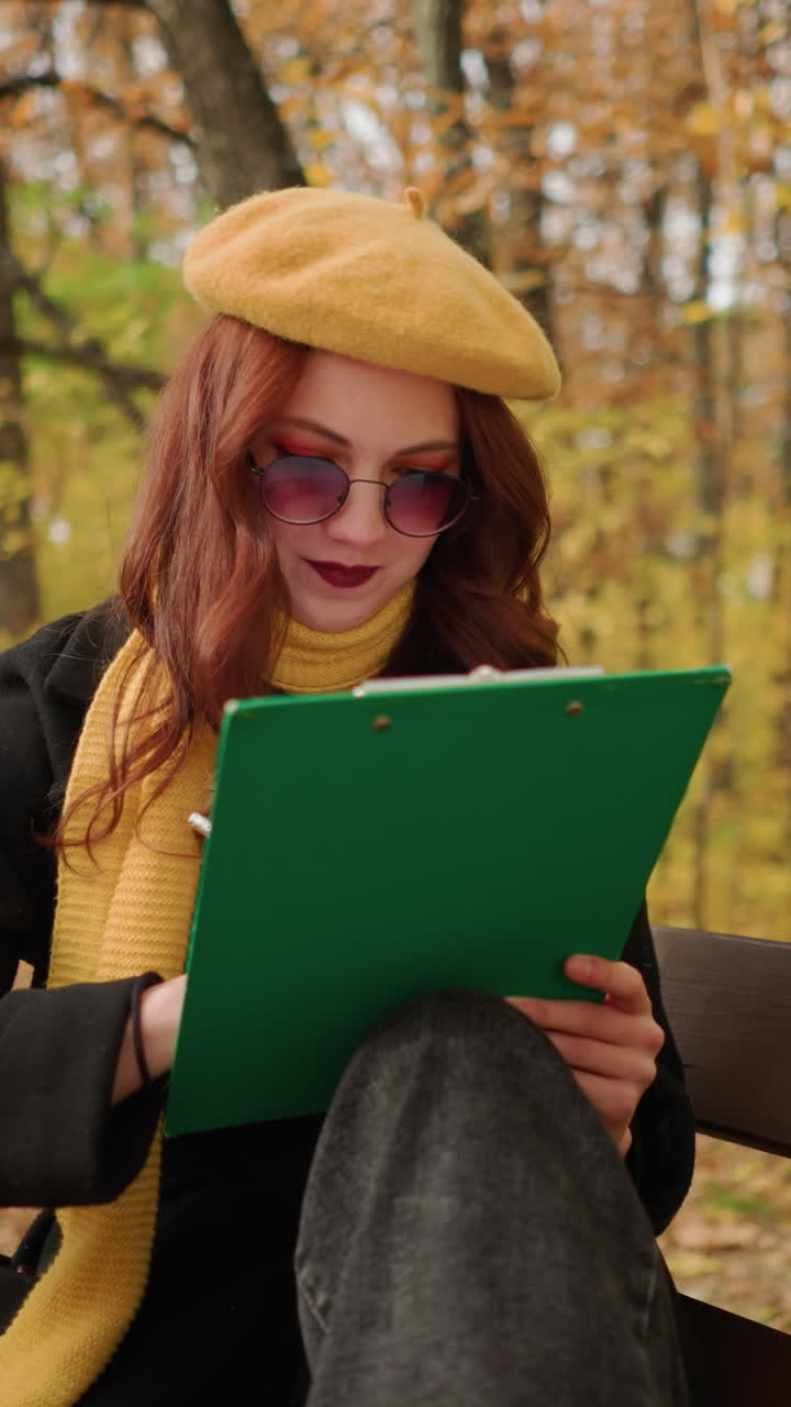 artista dibujando pensativamente en lienzo mientras está sentado en un banco de madera en un parque en un entorno tranquilo, rodeado de follaje de otoño, vestido con una elegante boina amarilla y bufanda