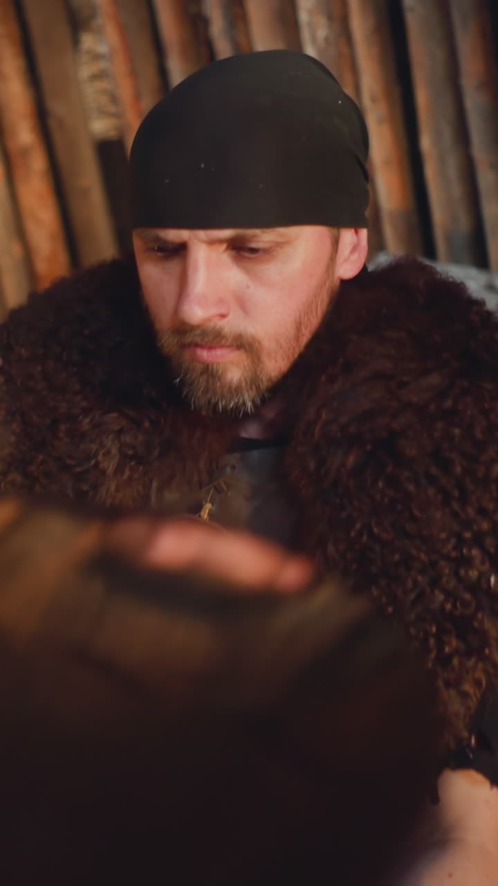 Serious bearded man with fur cover plays drums in ail house closeup. Folk rock band performs ritual Siberian music. Shamanic melodies