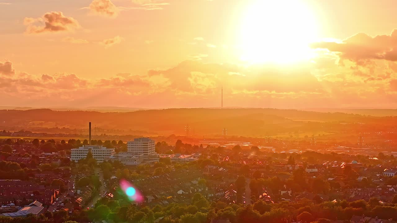 Static drone shot of Barnsley skyline at sunset with Barnsley Hospital in the foreground and the tall Emley Moor TV transmitter silhouetted on the horizon, warm golden tones cast across rooftops,