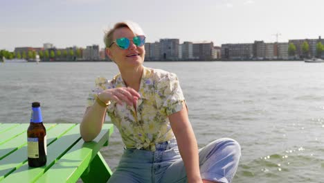 fun young woman enjoys the summer sun with a beer, sitting on picnic table, river and city in the background