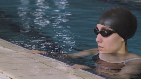 primer plano de una joven nadadora con gorra y gafas sumergiéndose en el agua y descansando en el borde de la piscina cubierta después de una sesión de natación