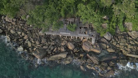 waves crash against a rocky shore where a decaying deck is nestled between boulders and overhanging trees.