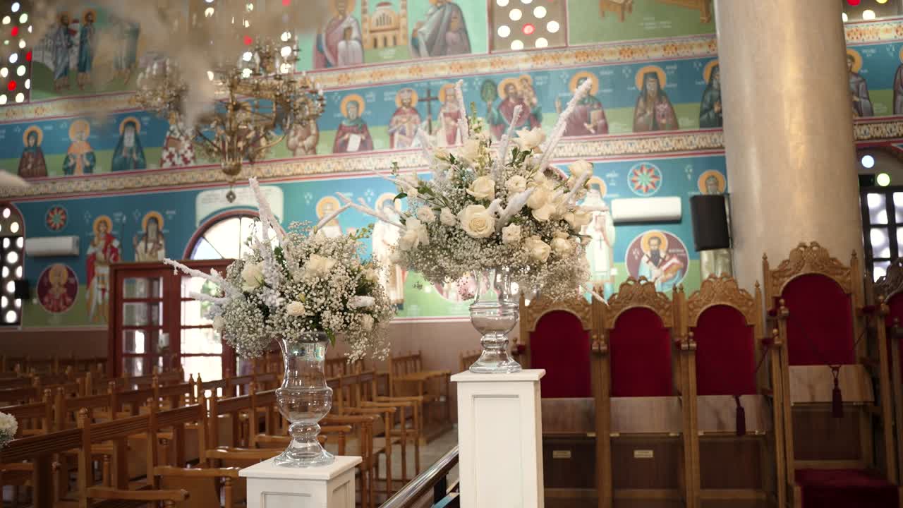 Guests gather for a wedding ceremony inside a church decorated with flowers. The interior features colorful murals and wooden chairs. The atmosphere is joyful as the event unfolds