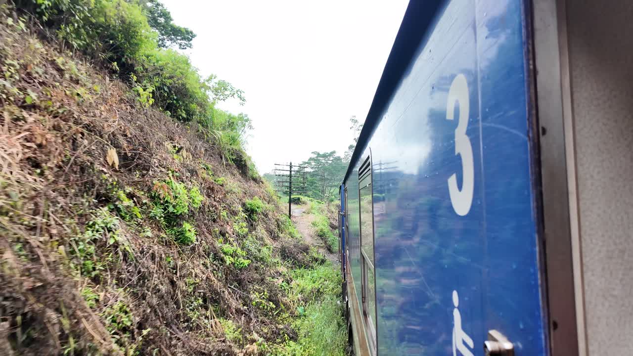 Blue train winding through a lush hillside landscape, with visible number three on the carriage, surrounded by greenery and an old telegraph pole nearby. Captivating travel scene.