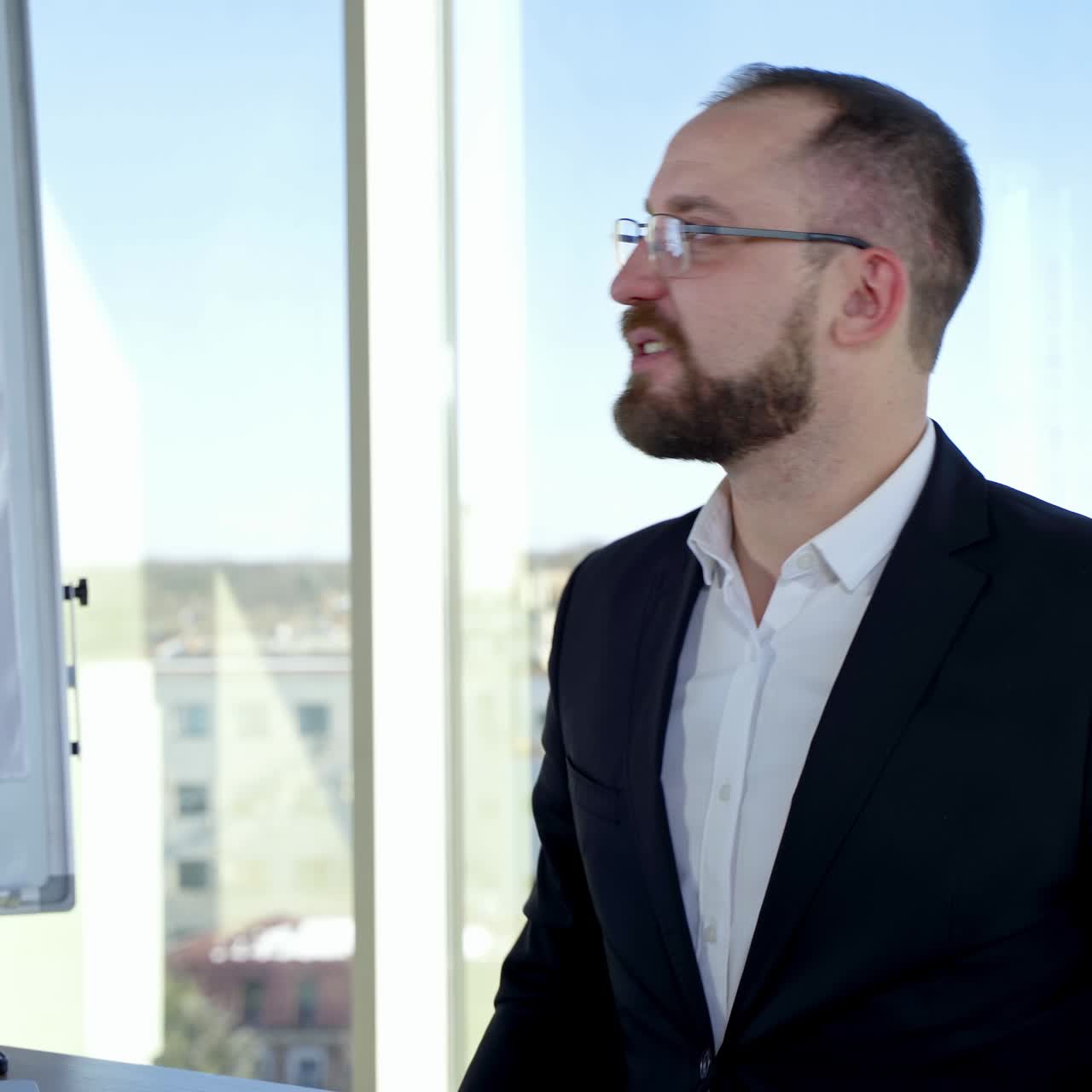 Smiling business worker talking to someone in office. Portrait of entrepreneur in glasses and costume sitting at his desk on the background of window in the city
