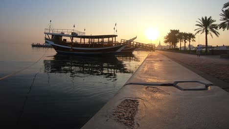 Landschaftlich-Ruhiger-Statischer-Blick-Auf-Katarer,-Die-Während-Des-Sonnenaufgangs-Am-Frühen-Morgen-Auf-Dem-Bürgersteig-Auf-Dem-Seeweg-Laufen,-Wobei-Das-Traditionelle-Dhow-Boot-Verankert-Ist