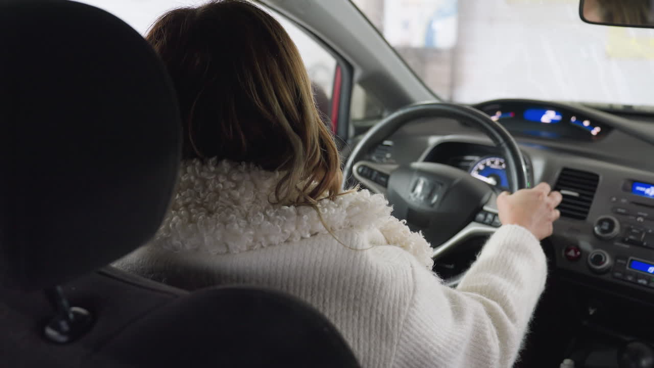 woman in white sweater sitting behind steering wheel inside car preparing to drive out from garage, hand resting on wheel, focus on dashboard controls, interior illuminated by soft daylight