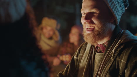 young-man-with-beard-chatting-to-woman-flirting-enjoying-romantic-connection-sitting-by-campfire-having-conversation-camping-with-friends