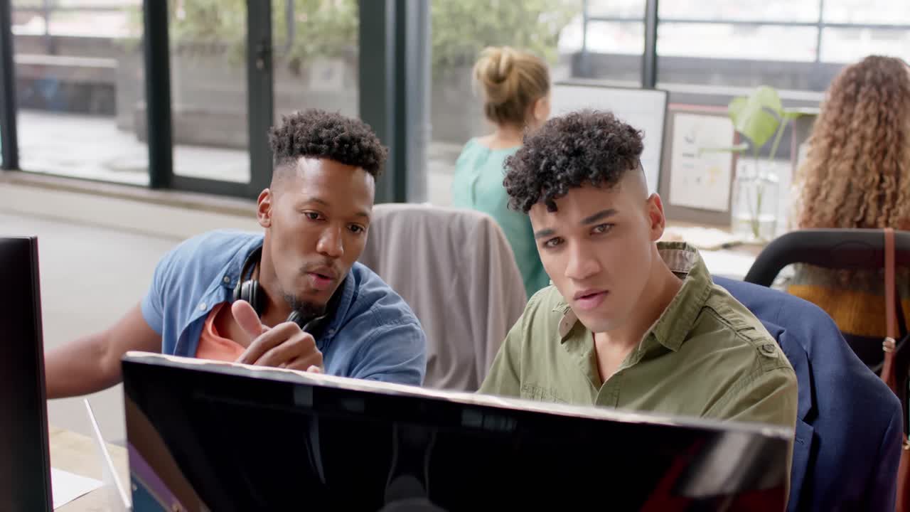 Two happy diverse male casual business colleagues in discussion at computer in office, slow motion