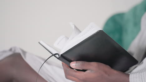 close-up of man in white bathrobe holding open black notebook with pages flipping, soft light highlighting fingers and pages, green velvet headboard in background, peaceful indoor atmosphere
