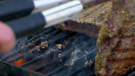 un par de pinzas para carne colocan un jugoso filete de costilla casi cocido en una parrilla y lo deslizan un poco a cámara lenta