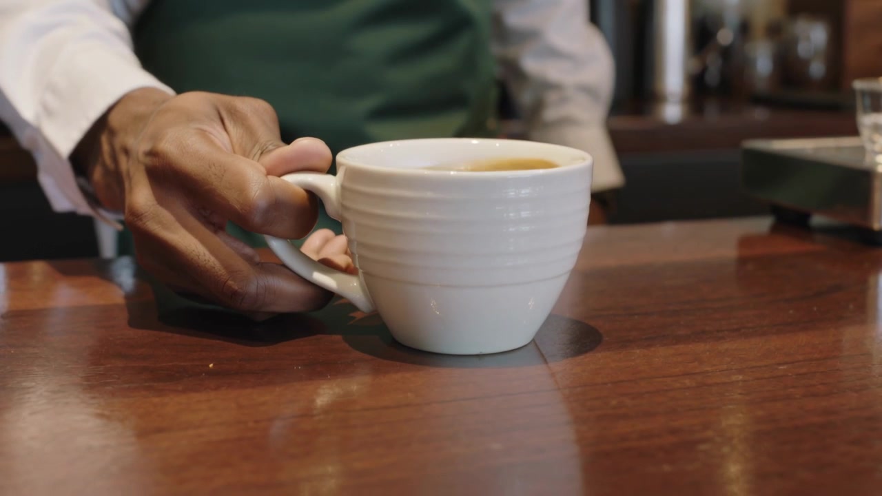 Barista con grembiule verde che si allunga, posizionando una grande tazza bianca sul bancone di servizio in legno, servendo un cliente.