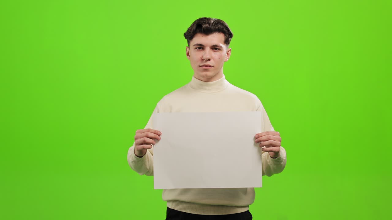 남자는 서서 카메라를 바라보고,  종이를 들고, 초록색 바탕에 서 있다.