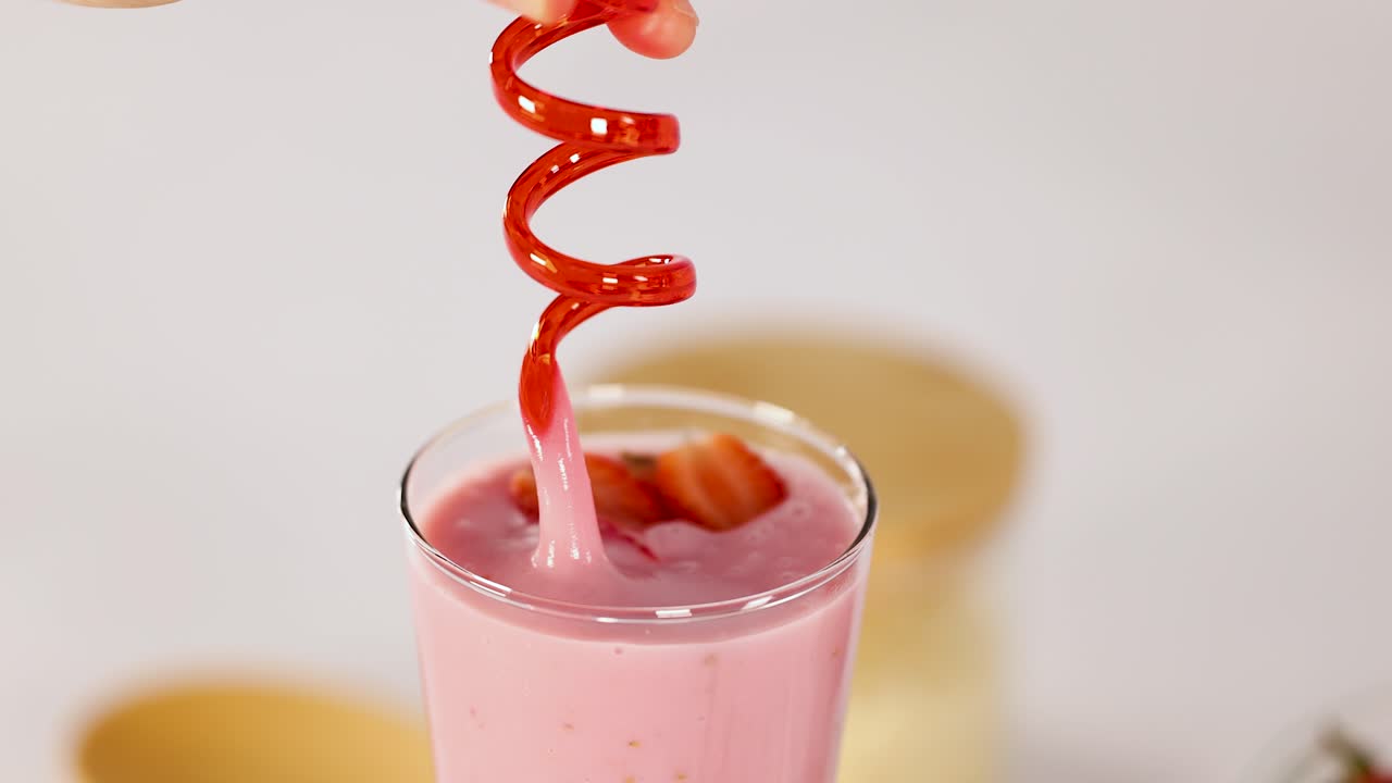 Hand inserts red spiral straw into creamy strawberry smoothie with fresh fruit garnish, bright lighting