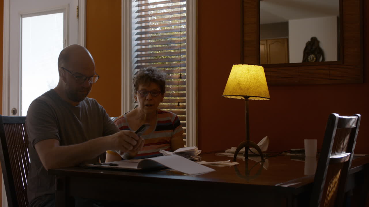 An adult man and his elderly mother sit together at a dining table in a warmly lit home, sorting through papers and bills. The son appears focused counting receipts, while the mother looks on. 24 fps.
