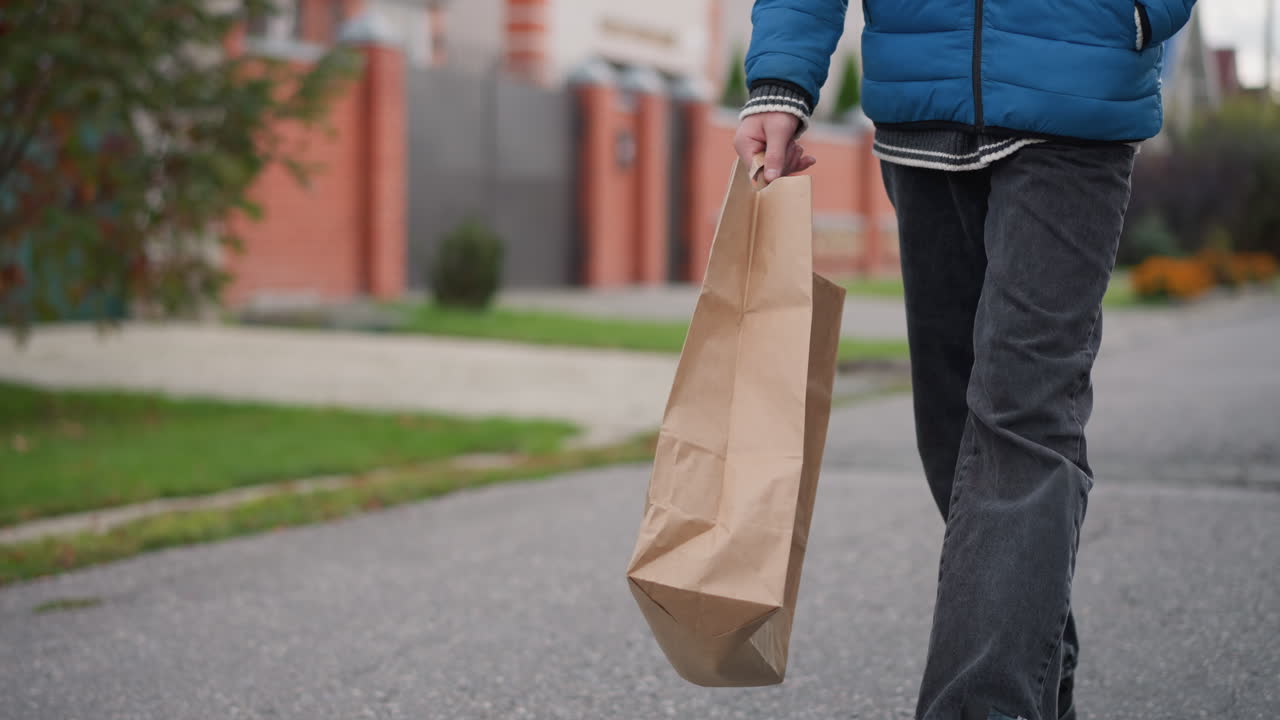 vista de ángulo inferior de un individuo con chaqueta azul y pantalones vaqueros caminando en un área residencial, llevando una bolsa de papel, árboles de otoño, casas, en el fondo con vegetación