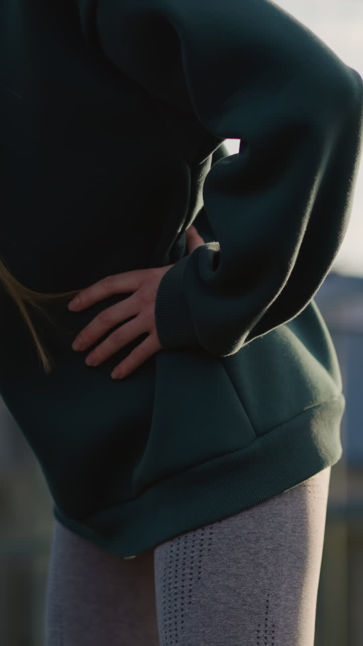 dama con sudadera verde y cabello largo realizando giros de cintura en la cancha al aire libre con el fondo borroso del edificio residencial y la red, capturando el estilo de vida saludable y la rutina de fitness