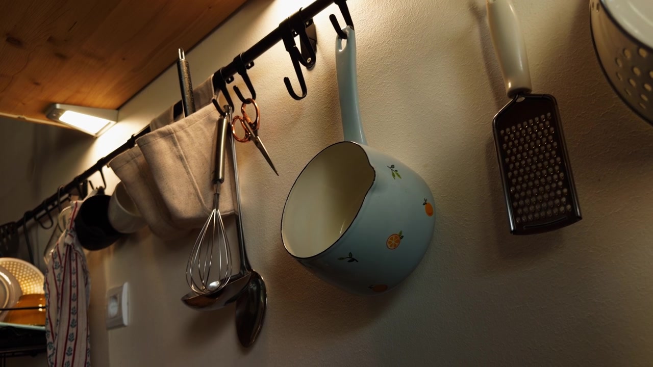 Saucepan, whisk, ladle, grater and other utensils hang from hooks on a kitchen wall, showing organized cookware and everyday home meal preparation.