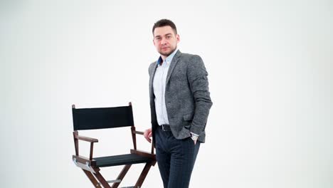 portrait of a young businessman in light studio. handsome man in elegant costume standing near the chair and looking at camera.
